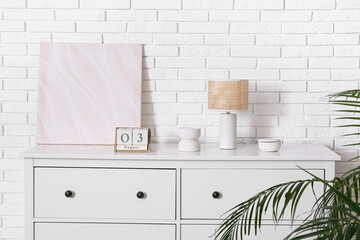 Cube calendar with date 3 AUGUST, lamp and painting on chest of drawers near white brick wall