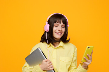 Young woman wearing pink headphones smiles while holding a laptop and a bright phone against a bold orange background, conveying playful multimedia study vibes.