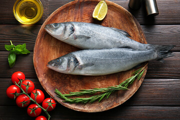 Plate with fresh raw seabass fishes and different ingredients on wooden background