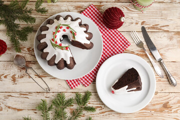 Plate with tasty cut Christmas cake, cutlery, fir tree branches, infuser and decor on light wooden background