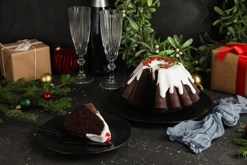 Plates with tasty cut Christmas cake, glasses, fir tree branches, gift boxes and decor on black grunge background