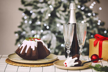 Tasty cut Christmas cake with bottle of champagne, glasses and gift box on table against blurred lights