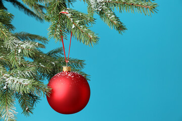 Red Christmas ball hanging on fir tree branch against light blue background, closeup. Space for text