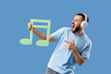 Singing young man in headphones pointing at paper music note on blue background