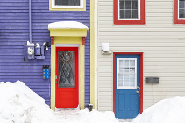 Two colorful doors on neighboring houses. There's a purple wooden house with a red door and glass windows. There's bright yellow trim around the door. The other house is white with a blue door.