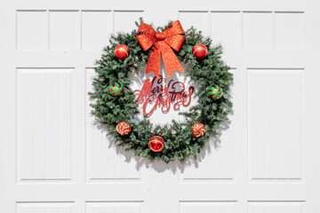 A lush green fir wreath hanging on a white wooden panel wall. The decoration has a large red bow with multiple red, green, and striped Christmas balls. Merry Christmas in red lettering in the middle.