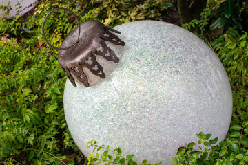 A large sparkly white round Christmas ornament made of plastic with a silver cap and loop. The holiday bulb is lying on lush green grass on a lawn. The festive bauble is illuminated in a white hue.