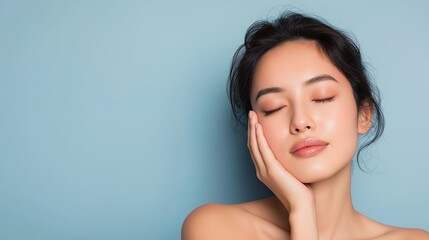 Serene Asian woman with flawless skin gently cradles her face against a blue backdrop, embodying natural beauty, calmness, and skincare wellness.