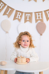 Adorable curly-haired toddler smiling with wooden birthday cake. Festive setup with balloons, paper banner, and cozy sweater at child party table