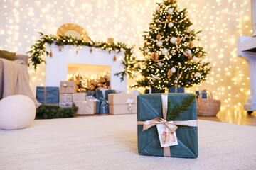 Christmas gift in green wrapping paper with ribbon on a carpet floor, with a blurred festive background of a decorated Christmas tree and shimmering lights.