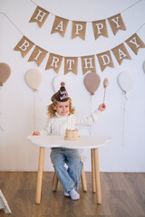 A cute caucasian child wearing a party hat sits at a small table with a mini cake for a first birthday celebration portrait.