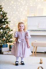Young caucasian girl standing next to Christmas tree with lights. Happy child enjoying holiday season. Festive childhood moment at home.