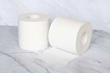 Two rolls of white toilet paper, with one partially unrolled, standing on a clean marble surface, symbolizing hygiene and household essentials.