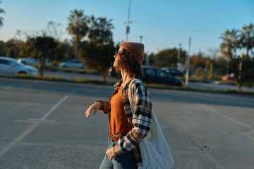 Woman walking in urban parking at golden hour, candid lifestyle portrait with beanie and knit bag,...