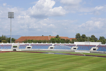 Calm, empty outdoor athletic stadium with green sport field and running track. blue grandstand seat waits for competition under partly cloudy sky background