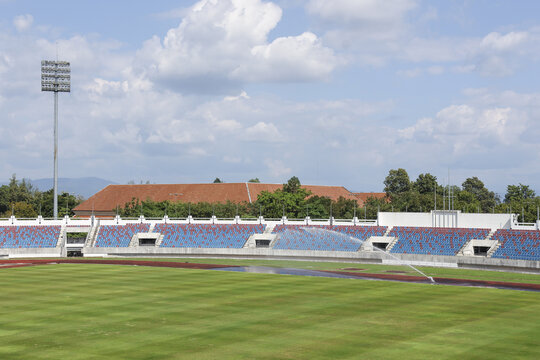 Calm, empty outdoor athletic stadium with green sport field and running track. blue grandstand seat waits for competition under partly cloudy sky background