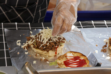 Chef preparing delicious burger with ground meat and cheese. This street food cooking process shows hand in glove carefully adding ketchup and other toppings