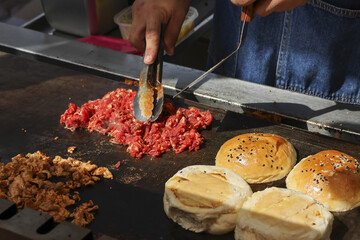 Focused chef cooking delicious street food, preparing beef burger on commercial grill. Chopped meat sizzling next to toasting sesame seed buns for fresh outdoor meal