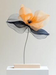 A delicate sculpture of a flower, crafted from translucent orange and black mesh fabric, is displayed on a simple wooden base.