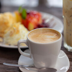 Hot coffee with latte art served in a white cup on wooden table, with blurred background of dessert and fruits. Perfect for breakfast themes, cafe promotions, lifestyle, and food blogs.