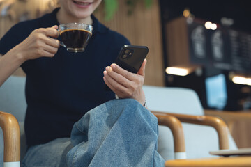 Happy young asian woman using smartphone and drinking coffee, social network at coffee shop. Smiling freelancer using mobile phone video call at cafe, modern people lifestyle