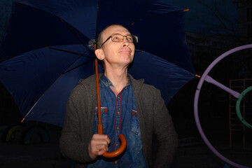 a man in glasses and a denim shirt holds a blue umbrella on a playground