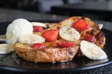 Sweet breakfast dish of golden French toast topped with banana slices, strawberries, vanilla ice cream, chia seeds, and syrup drizzle.
