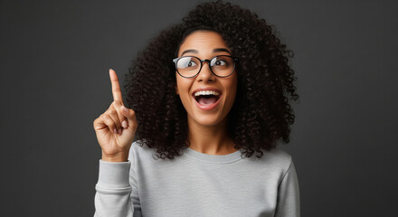 A joyful Indian woman with voluminous curly hair in a grey sweater raises one finger, suggesting an idea or guidance against a dark studio background.