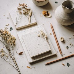 Two off-white speckled notebooks, tied with twine and adorned with dried flowers, rest on a light surface alongside pencils, a ceramic jug, and scattered botanicals.