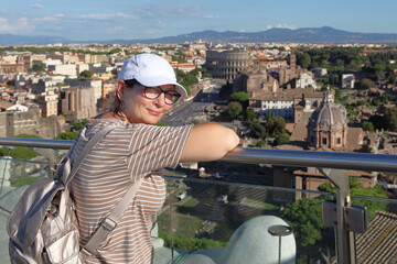 Tourist woman enjoying a panoramic view of the ancient Rome skyline from a high terrace, featuring the iconic Colosseum and Roman Forum ruins under a bright blue sky.