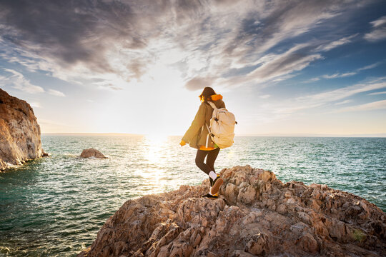 Young woman hiker with backpack is walking at sunset lake or sea beach against blue sky