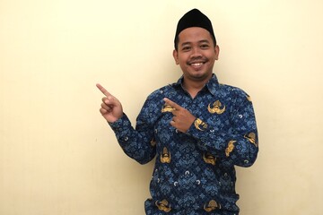 Happy Indonesian man in official PNS batik uniform and peci pointing to the side, isolated with copy space for advertisement or information.