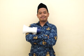 Indonesian civil servant wearing official PNS batik and peci standing confidently with arms crossed and megaphone, isolated with copy space for advertisement or leadership concept.