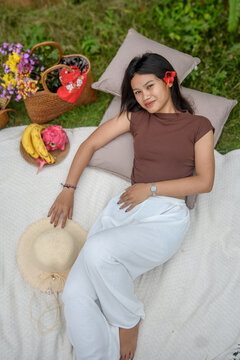 Top-down portrait of a young woman relaxing on a picnic blanket with fruits and flowers.