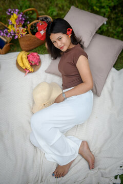 Top-down portrait of a young woman relaxing on a picnic blanket with fruits and flowers.