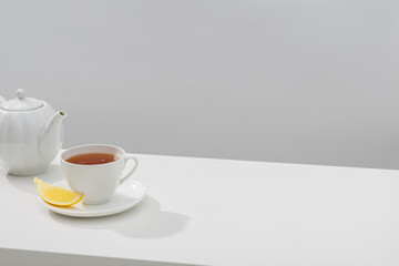 Fresh tea in a white ceramic cup, a lemon wedge, and a teapot on a white table. Light background.