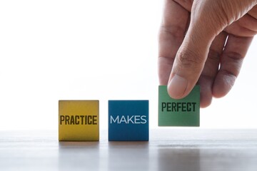A hand places the final wooden block to complete the motivational phrase, PRACTICE MAKES PERFECT. Skill development, training, dedication, improvement, and learning concept.
