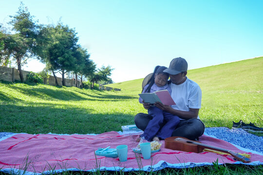 Man and Young Girl Sitting on Red and White Checkered Blanket Reading Book Together