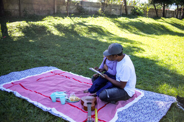 Father and Son Sit on Picnic Blanket Looking at Tablet with Ukulele and Snacks Nearby
