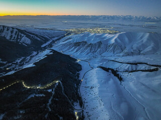 Wide aerial view of snow-covered mountains framing a valley with a glowing city, captured during the orange and blue hues of dawn or dusk