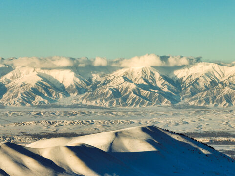 Telephoto aerial view of a vast, undulating snow-covered plain or valley floor with a massive, rugged, snow-capped mountain range visible on the distant horizon under a cloudy sky