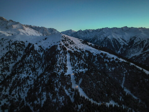 Aerial view of snow-covered mountain slopes at blue hour, featuring an illuminated ski run cutting through the dense, dark coniferous forest below the silent, snowy peaks