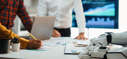 Professionals engage in a collaborative meeting with a humanoid robot, analyzing financial charts and data in a sleek, modern office setting.Noogenesis