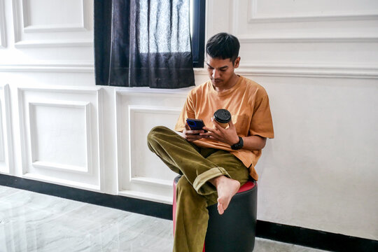 Young Man Casually Scrolling on His Smartphone While Holding Coffee Cup