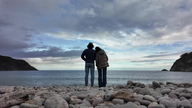 A couple stands on a rocky shoreline facing the sea under a dramatic cloudy sky.
