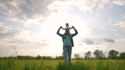 Father carries boy on shoulders walking slowly across tall summer field under soft sky. Father grips child hands gently. Peaceful green field and sky surround walk as father supports small child.
