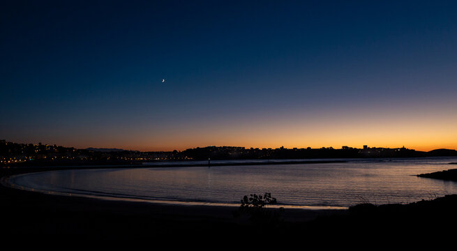 Blue hour over the bay of Cuchia with crescent moon and city lights, Cantabria, Spain