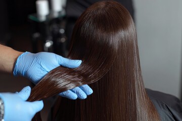 Fototapeta premium Glossy brown straightened hair is held in bluegloved hands Focus is on the hair