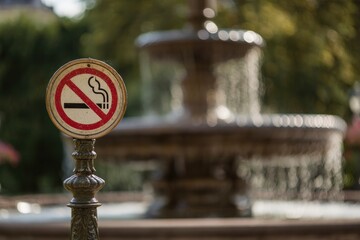 A No Smoking sign stands before a blurred fountain with greenery in the background