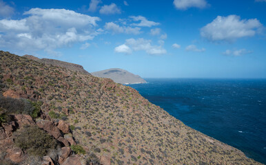 A coastal view from the Amatista viewpoint, Almeria, Spain. Rugged hills with sparse greenery, a deep blue sea, and distant mountains beneath a mostly clear sky with a few clouds